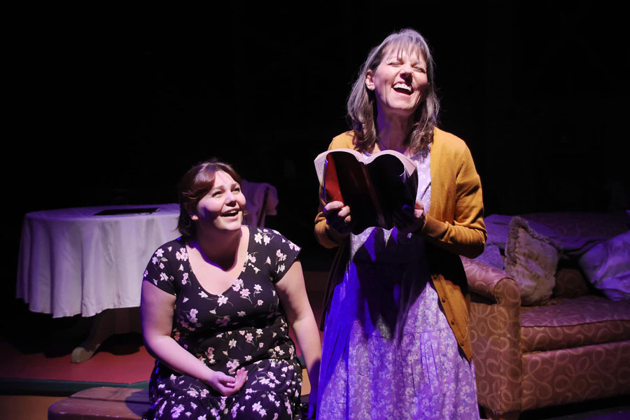 Two ladies on stage laughing while one is reading a book.